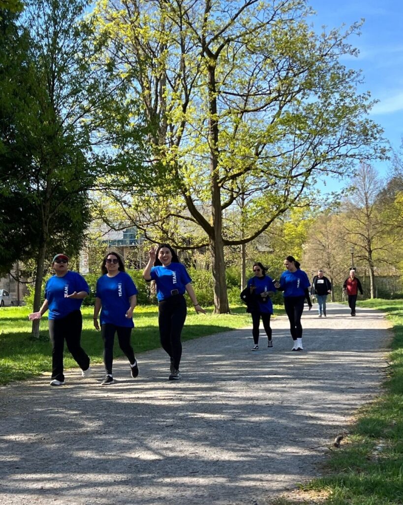 Group of six people in blue shirts walking together on a sunny park path among trees.