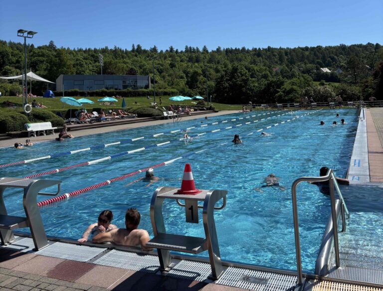 Outdoor public swimming pool with lane dividers, swimmers in the water, poolside loungers and turquoise umbrellas, and a grassy, tree-filled background.